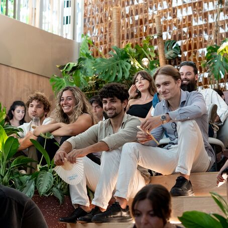 Audience seated on steps attending indoor event surrounded by lush plants